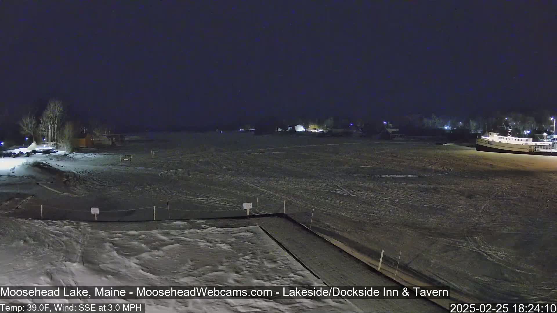 A nighttime view of a snow-covered lakefront with some buildings and a boat, under a dark sky with a 3.0 mph SSE wind and a temperature of 39°F.