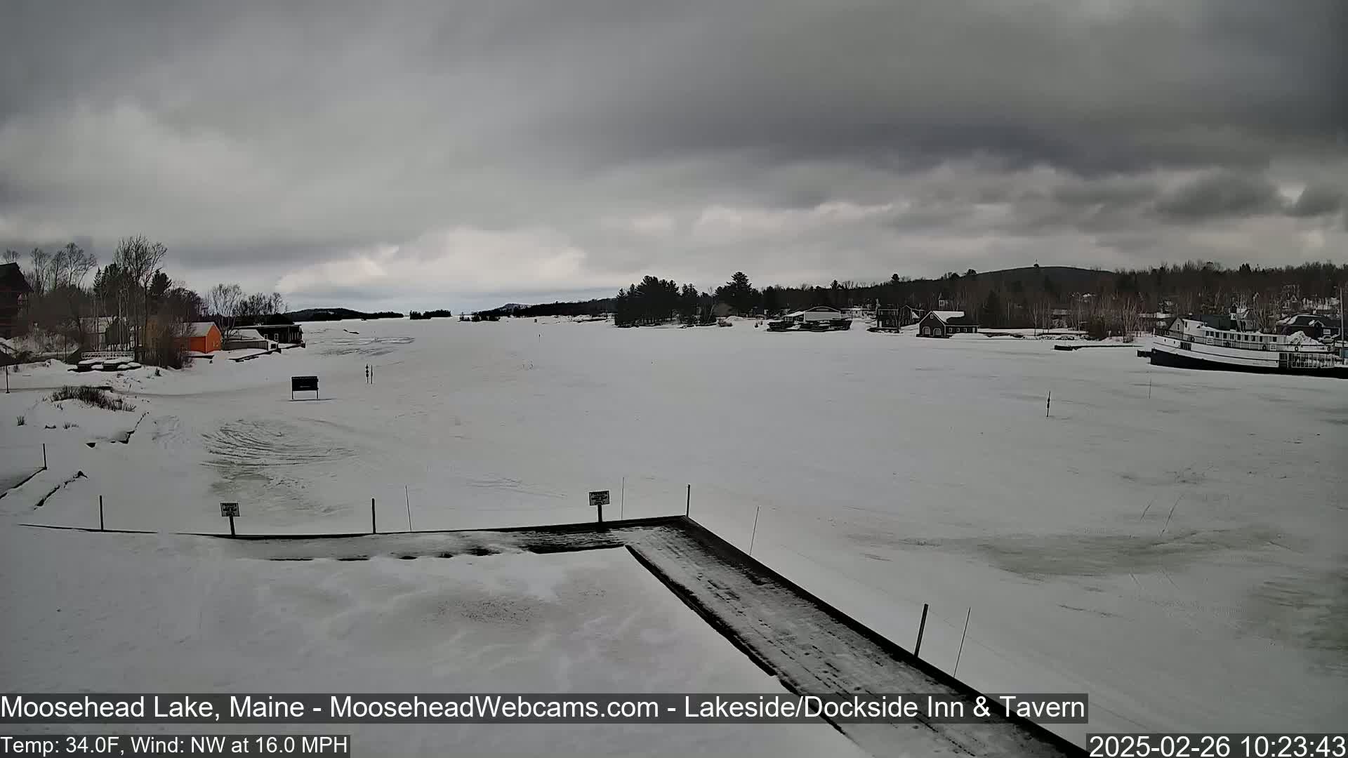 A frozen lake, under an overcast sky, is seen from a snowy shoreline with a few buildings and a boat visible in the distance.