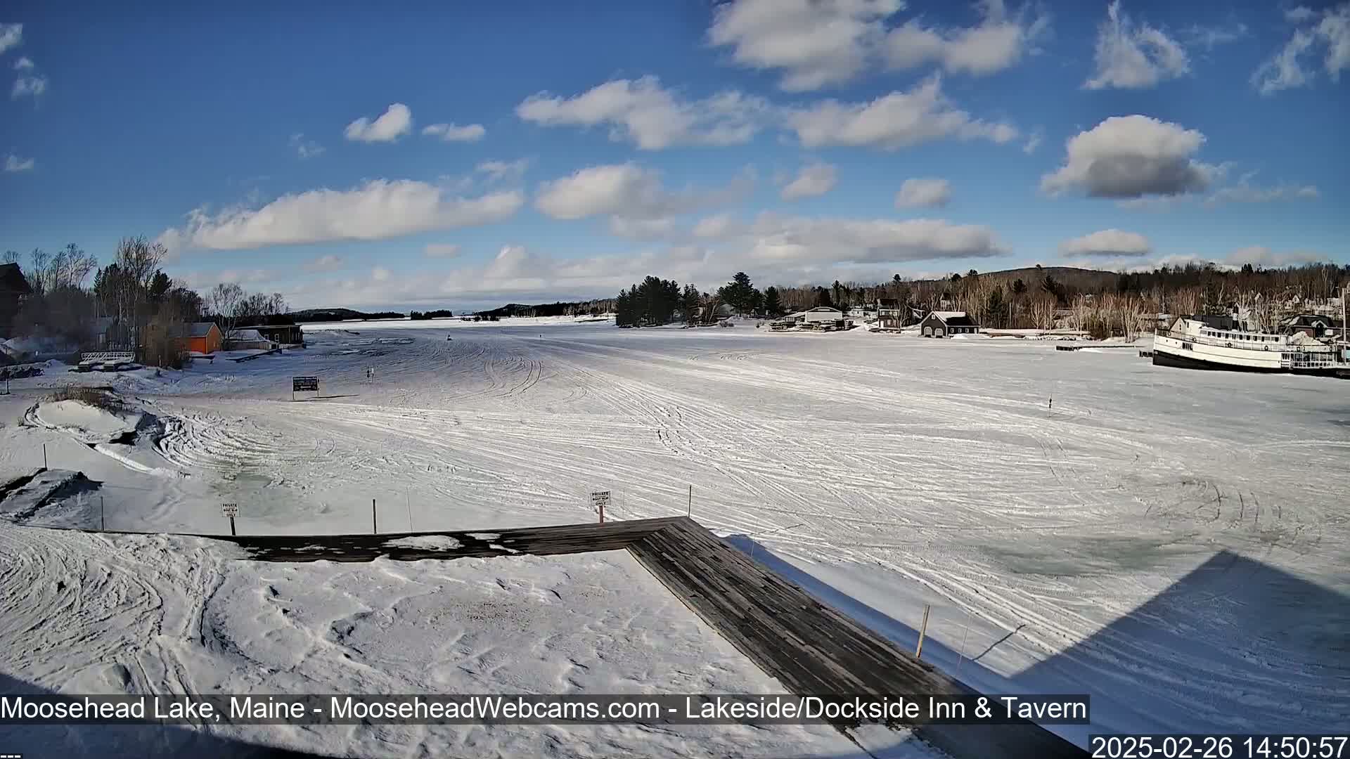 A snow-covered frozen lake with a wooden dock in the foreground, under a partly sunny sky, features buildings and a boat on the far side.