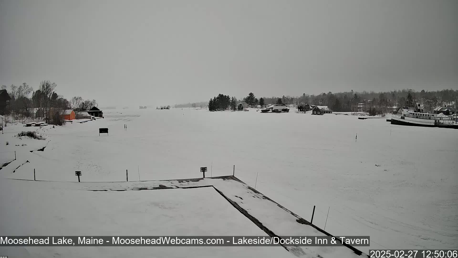 A snow-covered frozen lake is seen from a snow-covered dock, with a few buildings and a boat visible in the distance under an overcast sky.