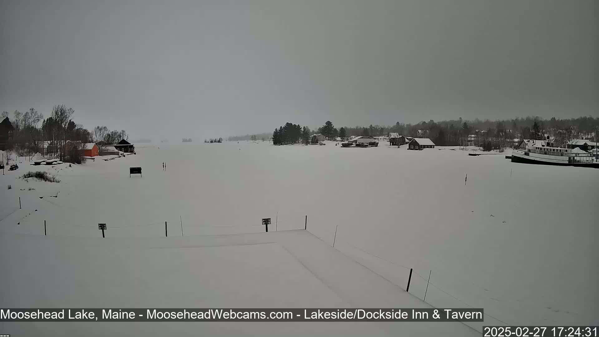 A snow-covered frozen lake, with a few buildings and trees visible on the shore, under an overcast sky.