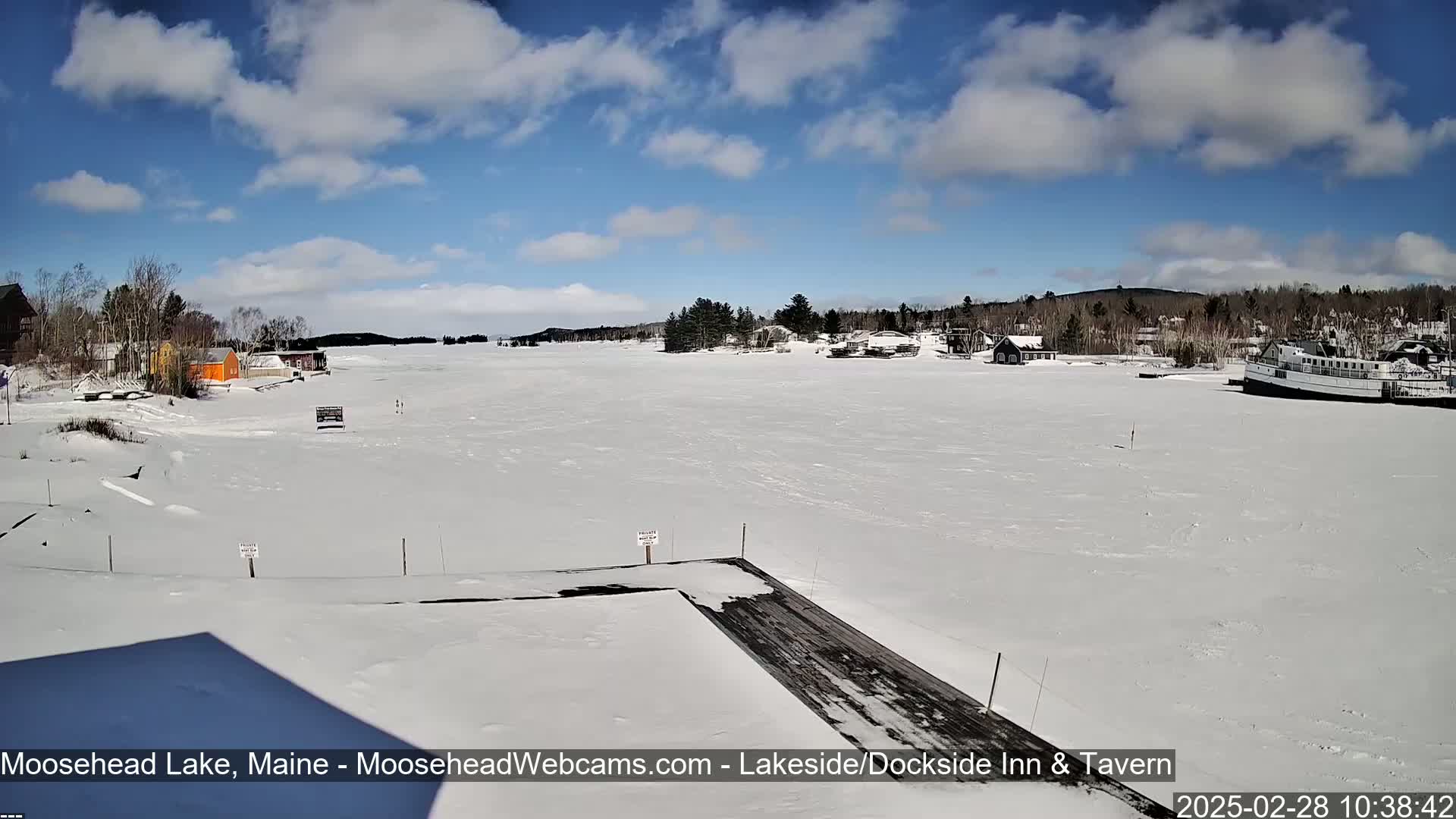 A snow-covered lake under a partly cloudy blue sky, with buildings and a boat visible in the distance.