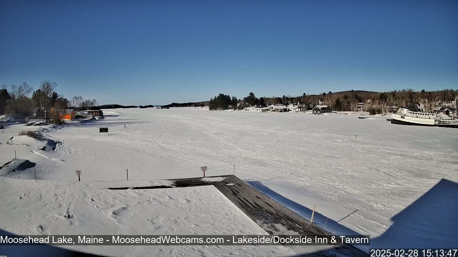 A frozen lake stretches out under a clear blue sky, with a few buildings and a large boat visible on the shore, surrounded by snow-covered land.