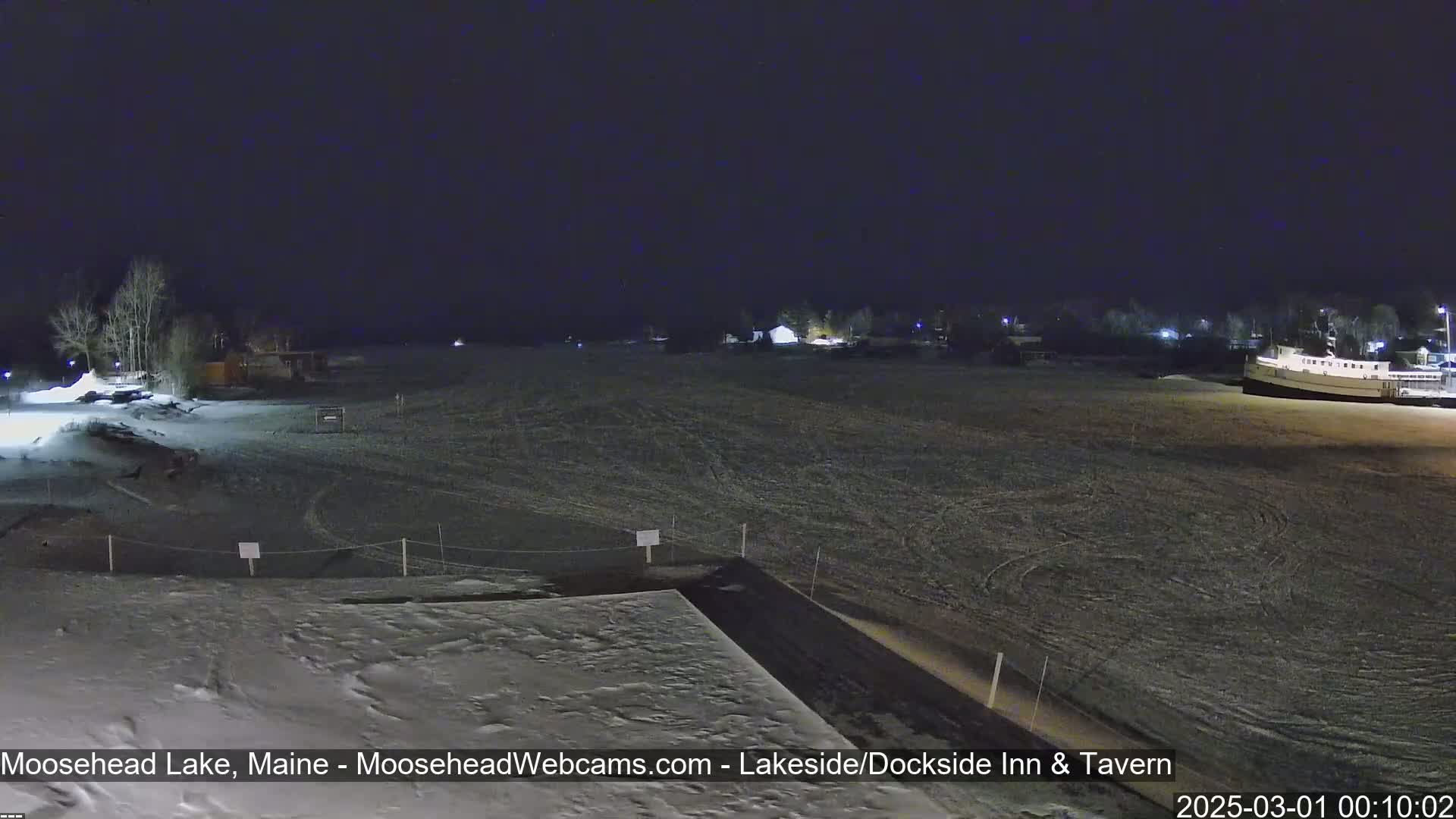 A snow-covered lakeside area at night, with a few buildings visible in the distance under a dark, clear sky.