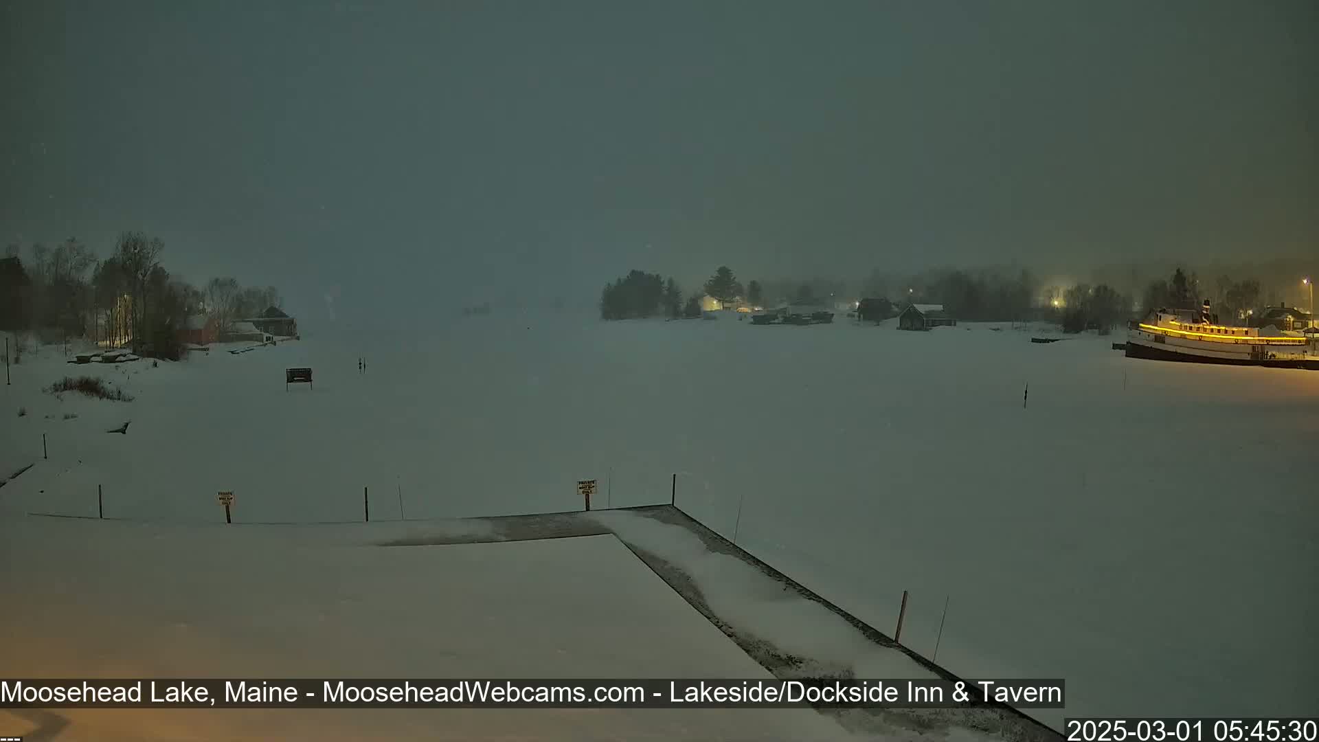 A snow-covered lake at night, with buildings and a boat visible in the distance under a dark, overcast sky.