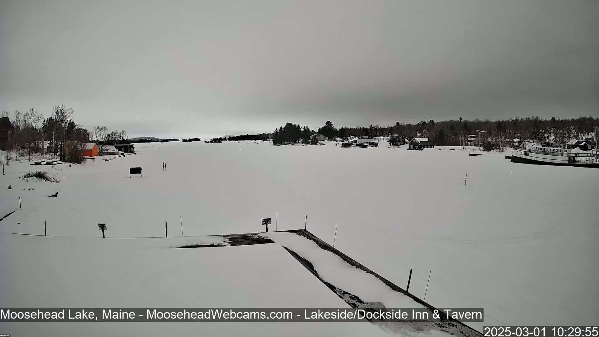 A snow-covered lake is seen from a snowy dock, with a few buildings and a boat visible in the distance under an overcast sky.