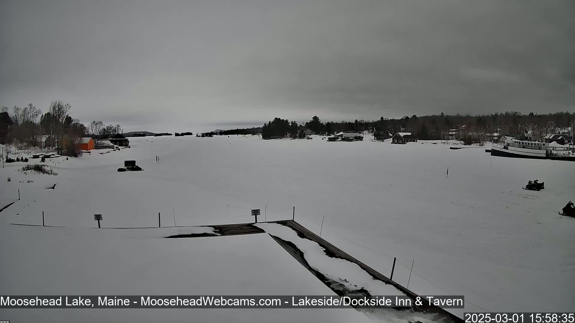 A snow-covered frozen lake with a few buildings and snowmobiles visible under an overcast sky.