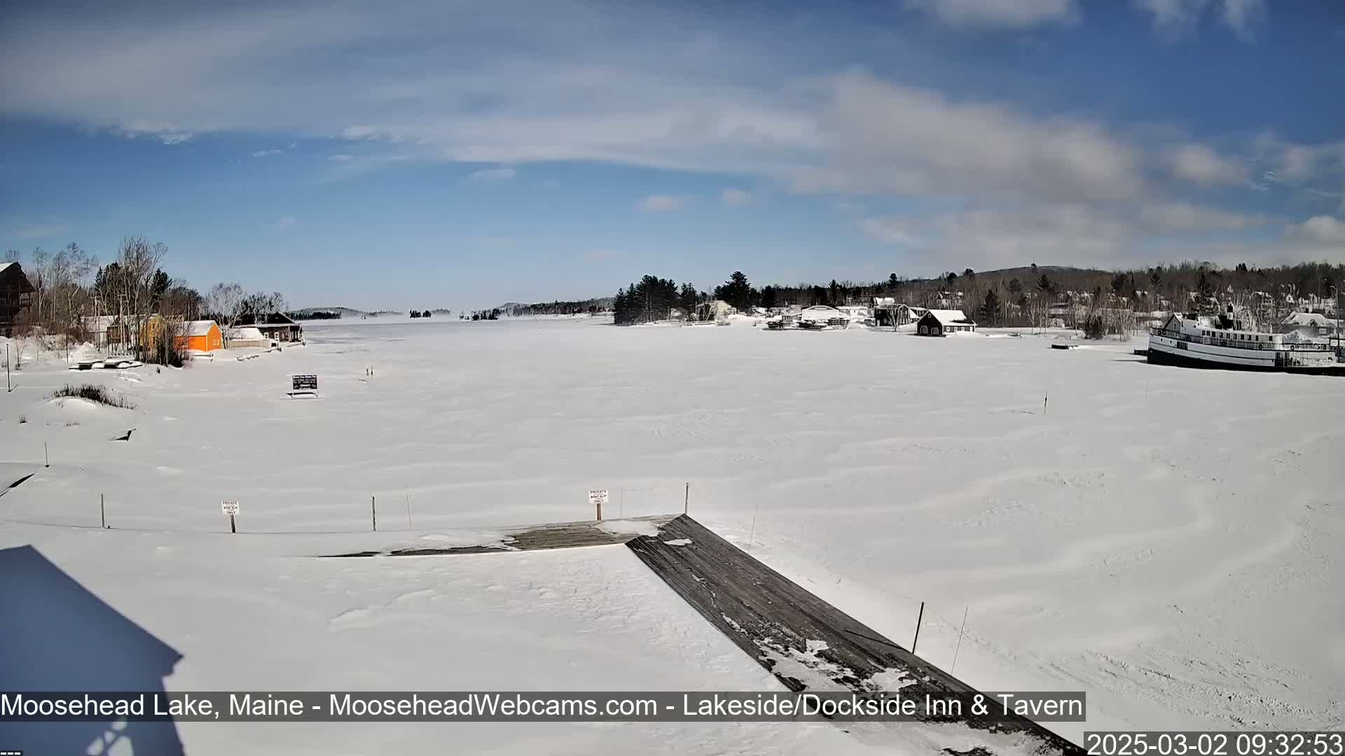 A snow-covered lake with a few buildings and a boat on the frozen water under a mostly sunny sky.