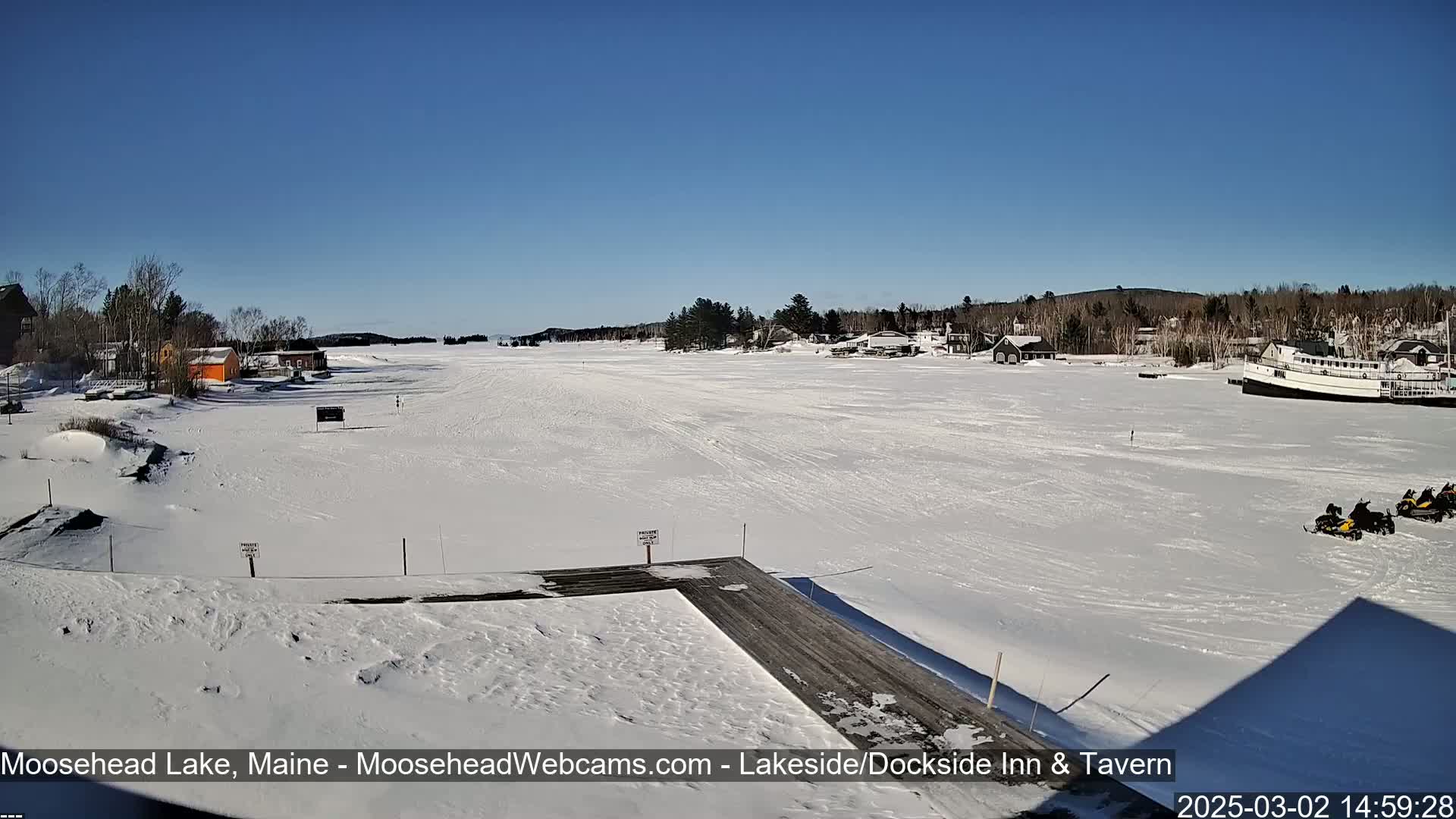 A snow-covered frozen lake with a few buildings and snowmobiles on the shore under a clear blue sky.