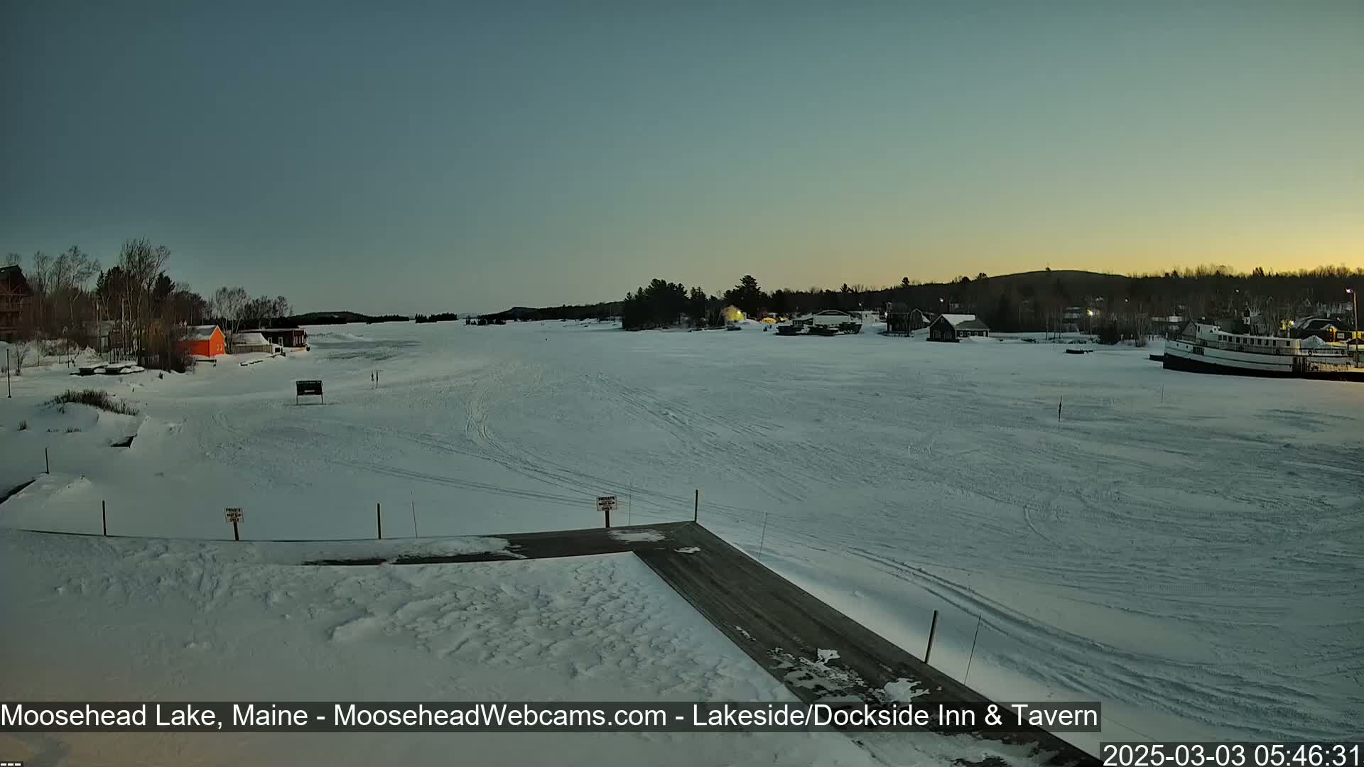 A frozen lake at dawn is seen from a snowy dock, with a few buildings and a boat visible in the distance.