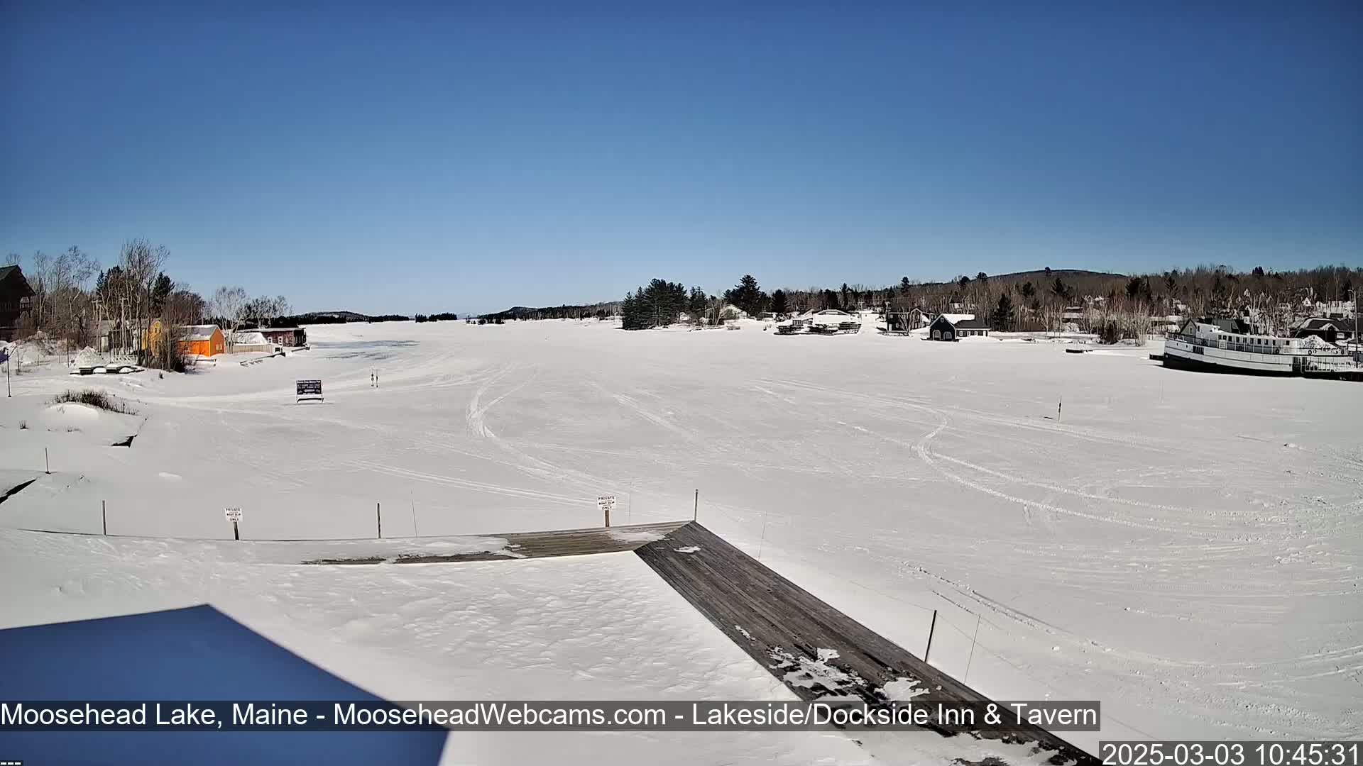 A snow-covered frozen lake stretches out under a clear blue sky, with buildings and a boat visible in the distance.