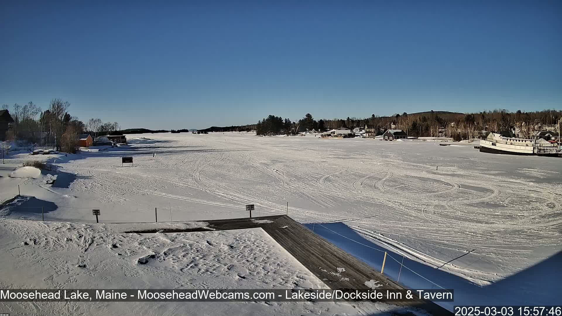 A frozen lake, under a clear blue sky, shows tire tracks on the ice and a few buildings in the distance.