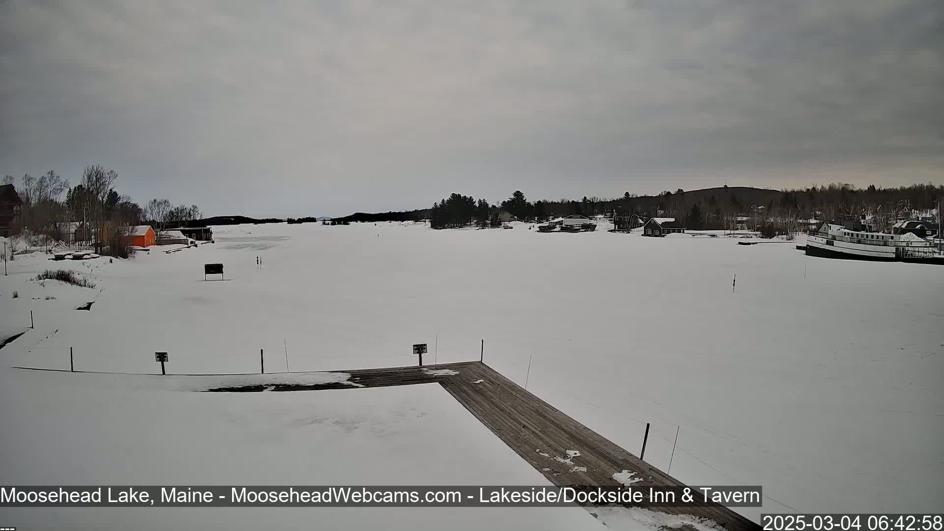 A snow-covered frozen lake is viewed from a wooden dock, with a few buildings and a boat visible in the distance under an overcast sky.