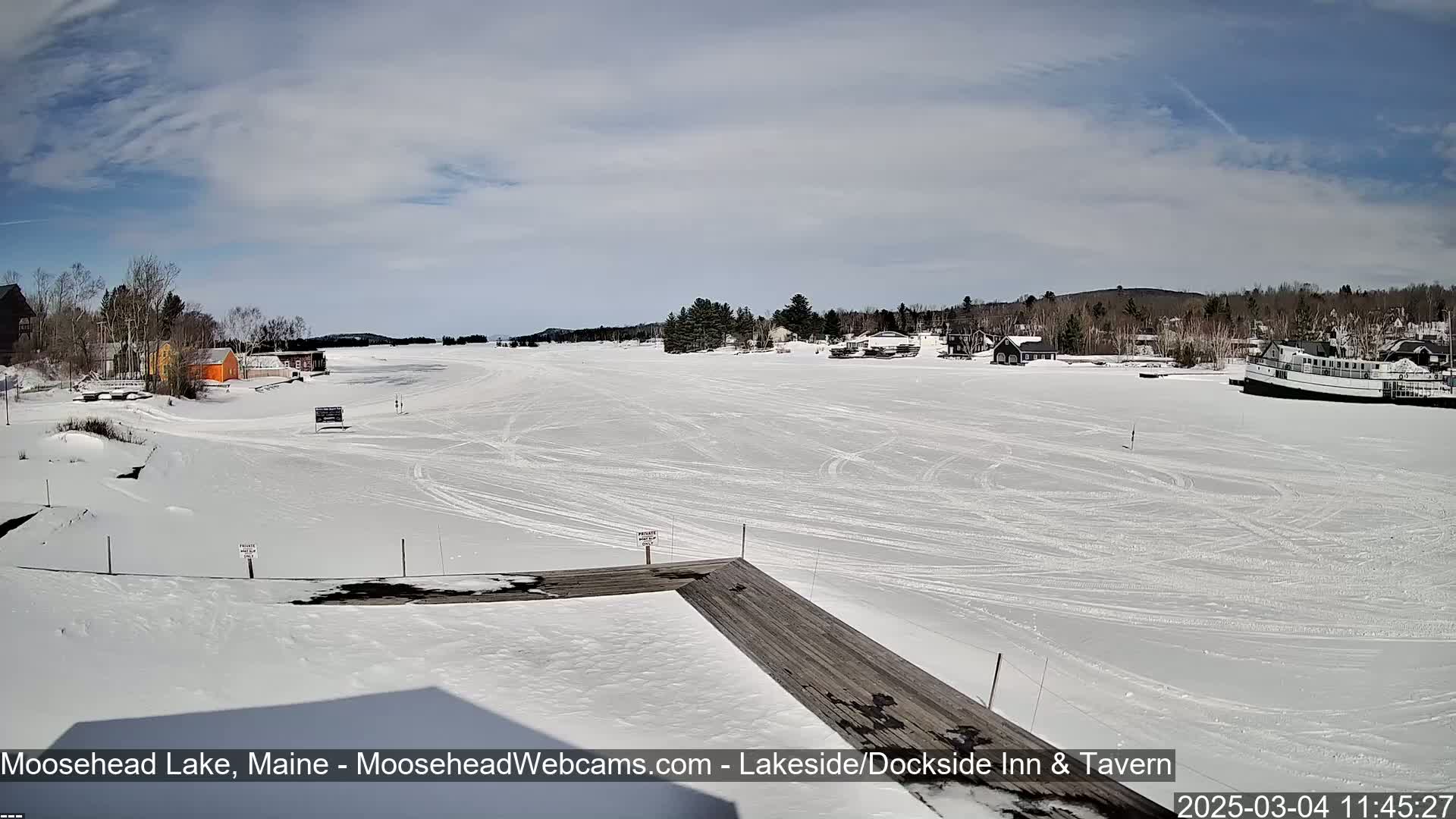 A snow-covered frozen lake stretches out from a wooden dock under a partly cloudy sky.