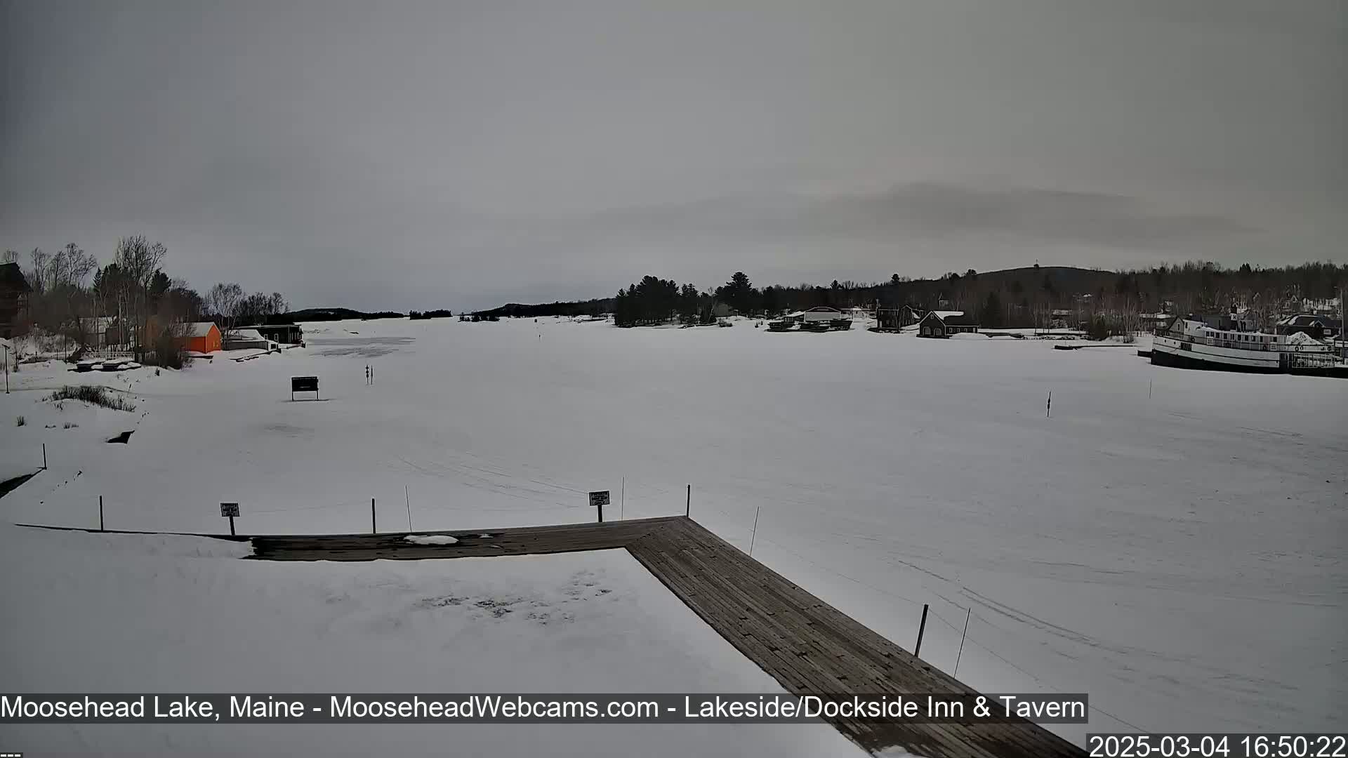 A snow-covered lake is viewed from a wooden dock, with a few buildings and a boat visible in the distance under an overcast sky.