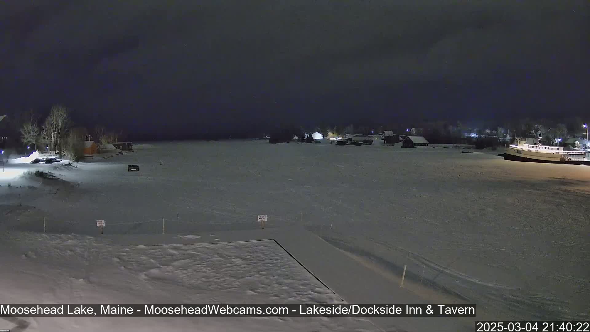 A snow-covered lake at night is seen from a snowy shoreline with some buildings and a boat in the distance under a dark sky.