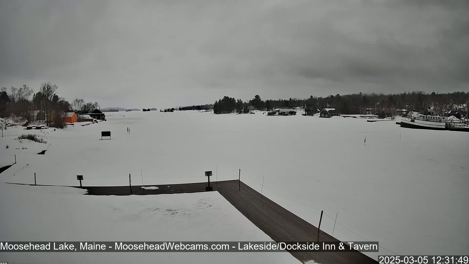 A snow-covered lake with a wooden dock in the foreground, a few buildings along the shore, and a boat in the distance under an overcast sky.