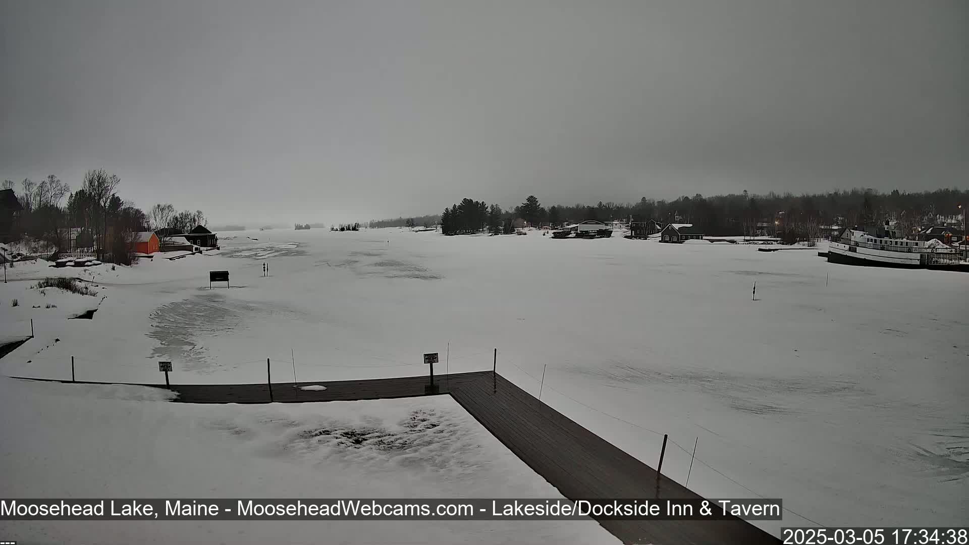 A snow-covered frozen lake is seen from a dock, with a few buildings and trees visible in the distance under an overcast sky.