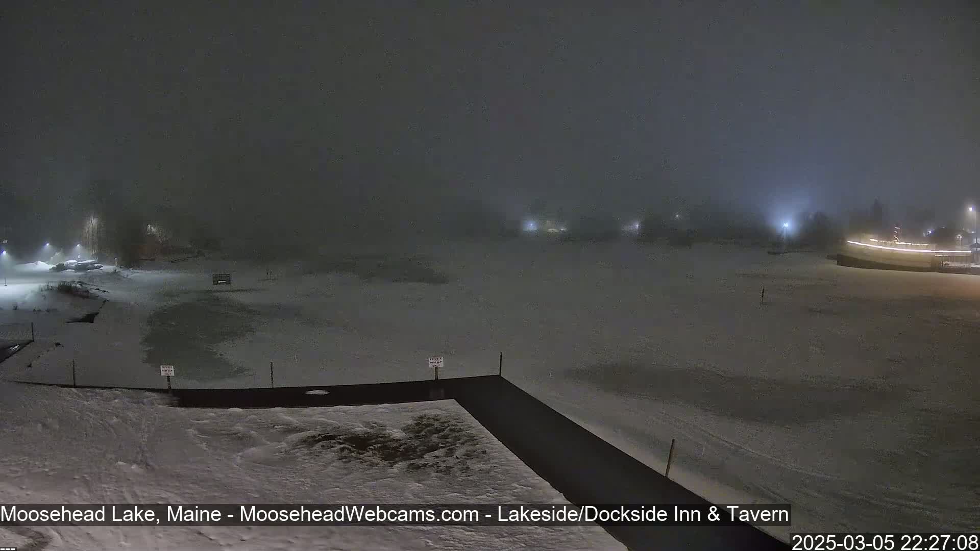 A snowy, foggy nighttime scene shows a partially frozen lake and a snow-covered shoreline with dimly lit buildings in the distance.