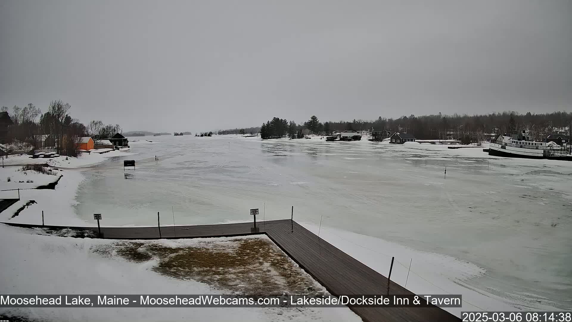 A partially frozen lake is seen from a snow-covered shoreline, with a wooden dock in the foreground and buildings and a boat visible in the distance under an overcast sky.