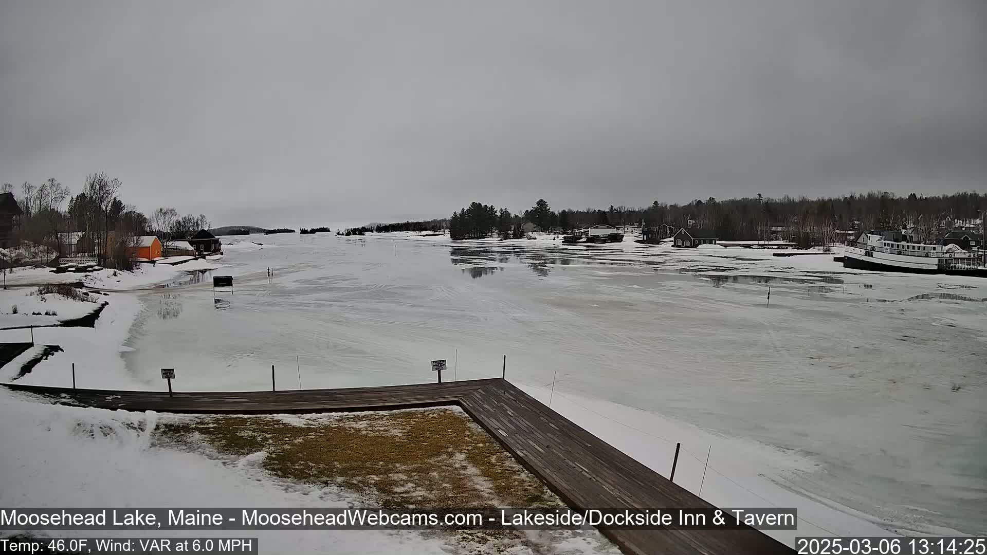 A partially frozen lake is seen from a snowy lakeside dock, with buildings and trees visible in the background under a gray sky.