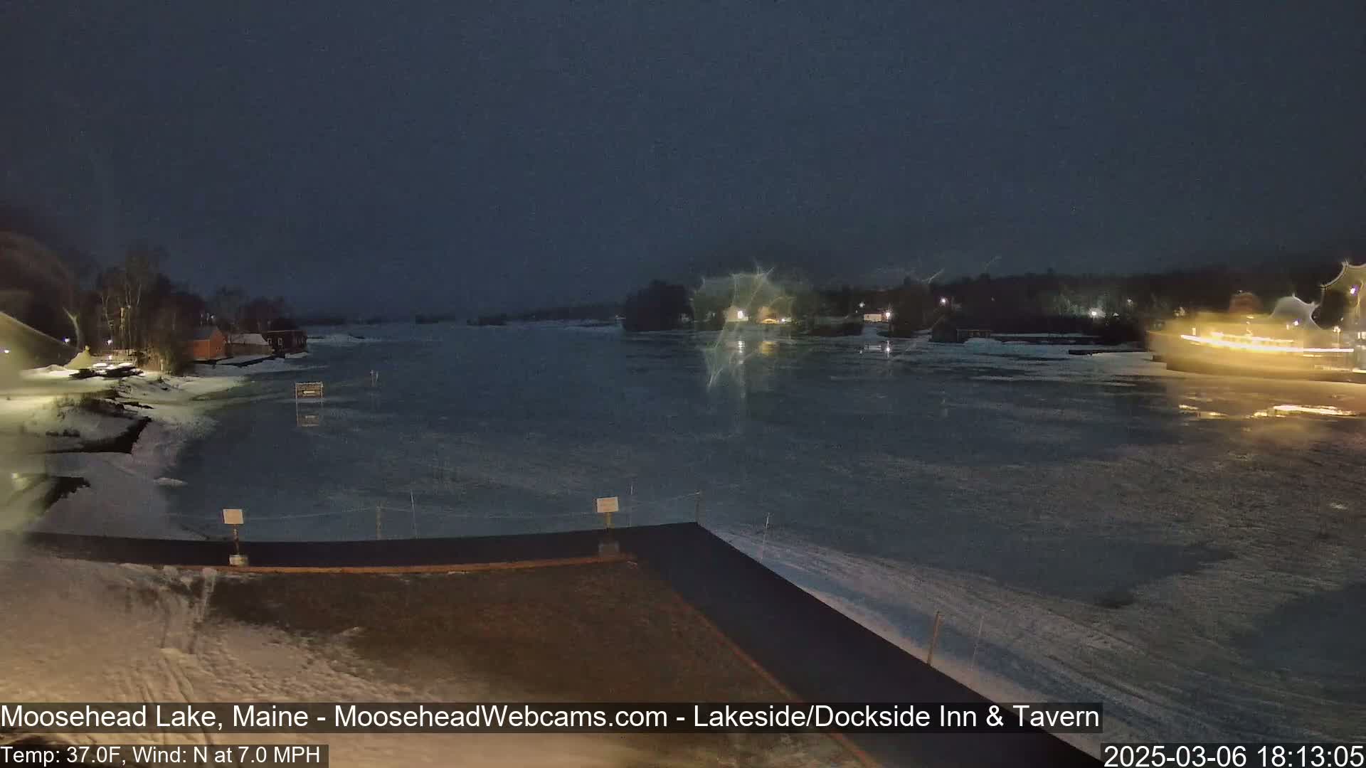 A partly frozen lake is visible at night under a dark sky, with a 7 mph north wind and a temperature of 37°F; buildings are lit up on the far shore.