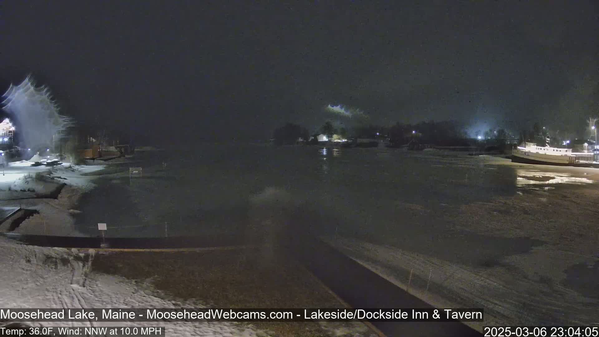 A nighttime view of a partly frozen lake with snow blowing intensely near the shore under a dark sky.