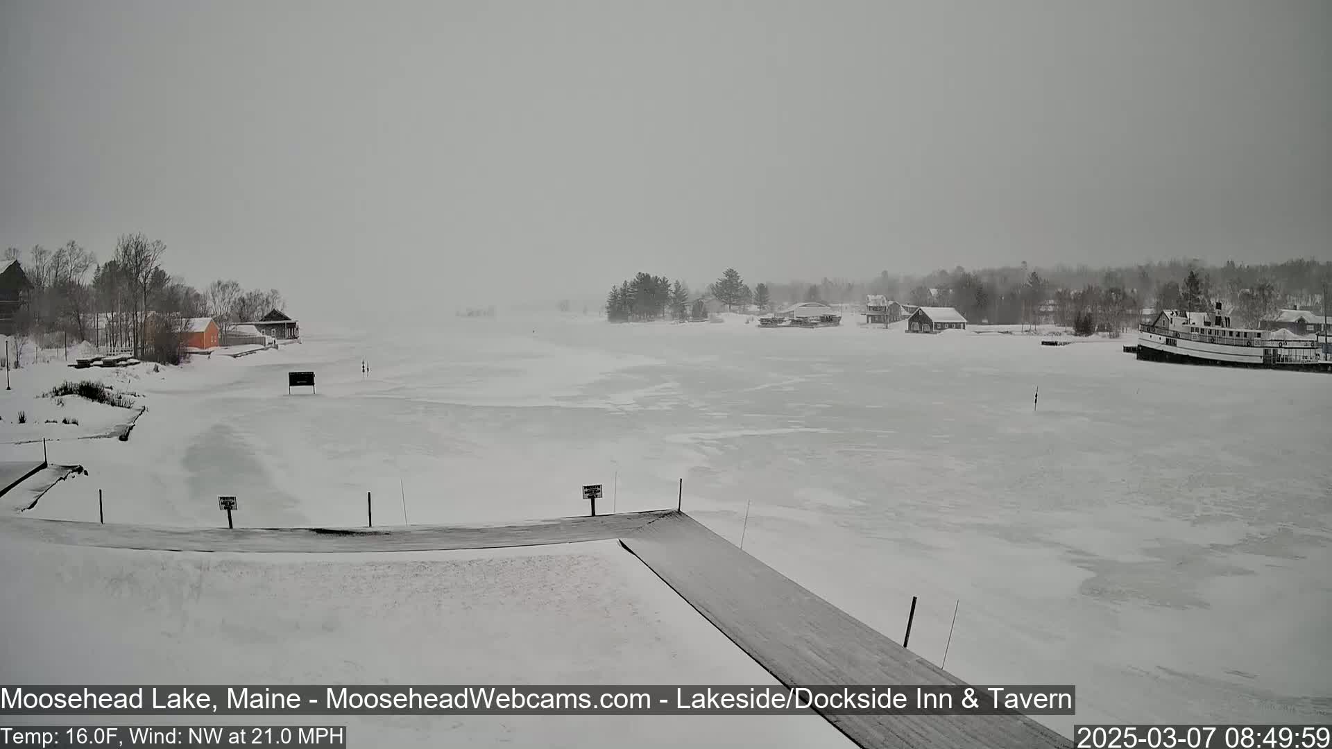 A snow-covered frozen lake is seen with a few buildings and a boat on the shore under an overcast sky, with a northwest wind blowing at 21 mph and a temperature of 16°F.