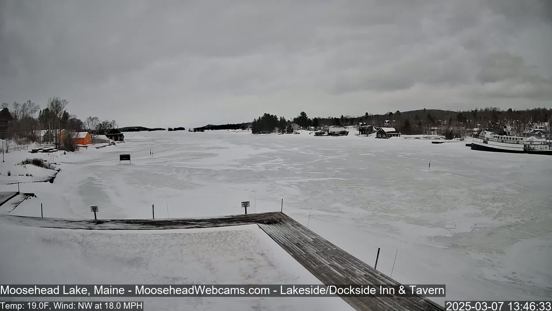 A mostly frozen lake is viewed from a snow-covered dock on an overcast day with a northwest wind.