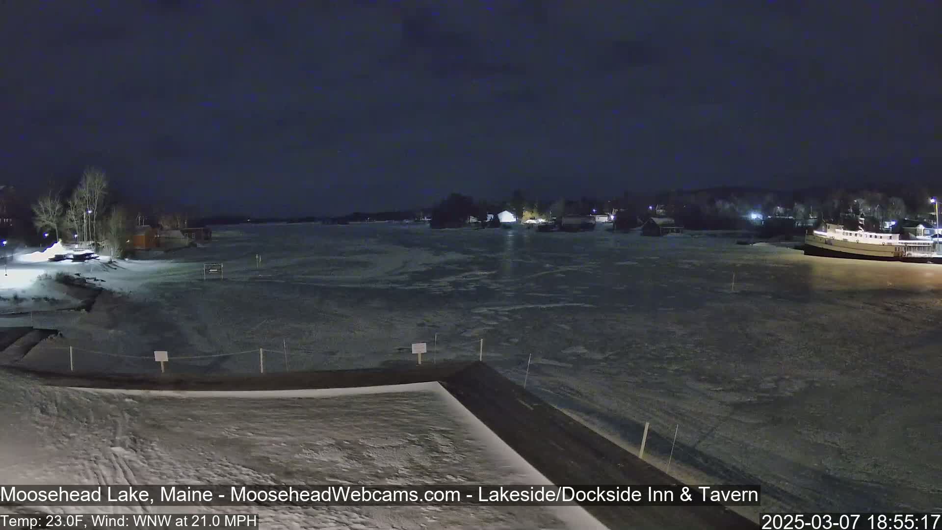 A partly frozen lake is visible at night under a dark sky, with buildings and a boat along the shore, and snow on the ground.