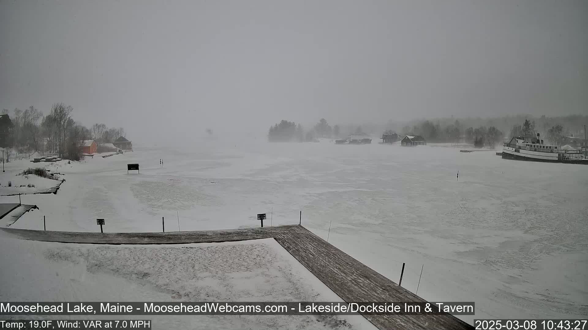 A snow-covered, partially frozen lake is seen from a snow-covered dock, with buildings and a boat visible in the distance during a snowstorm.