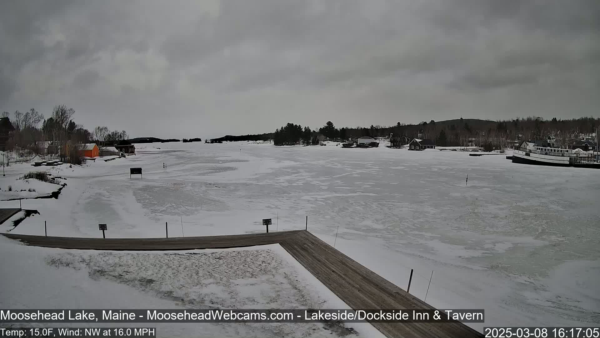 A mostly frozen lake is viewed from a snow-covered dock under an overcast sky.