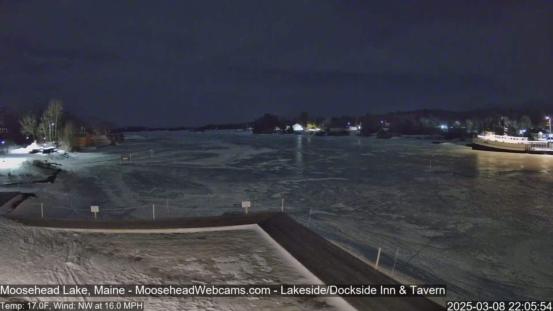 A partially frozen lake is viewed at night from a snow-covered dock under a dark sky, with a northwest wind at 16 mph and a temperature of 17 degrees Fahrenheit.