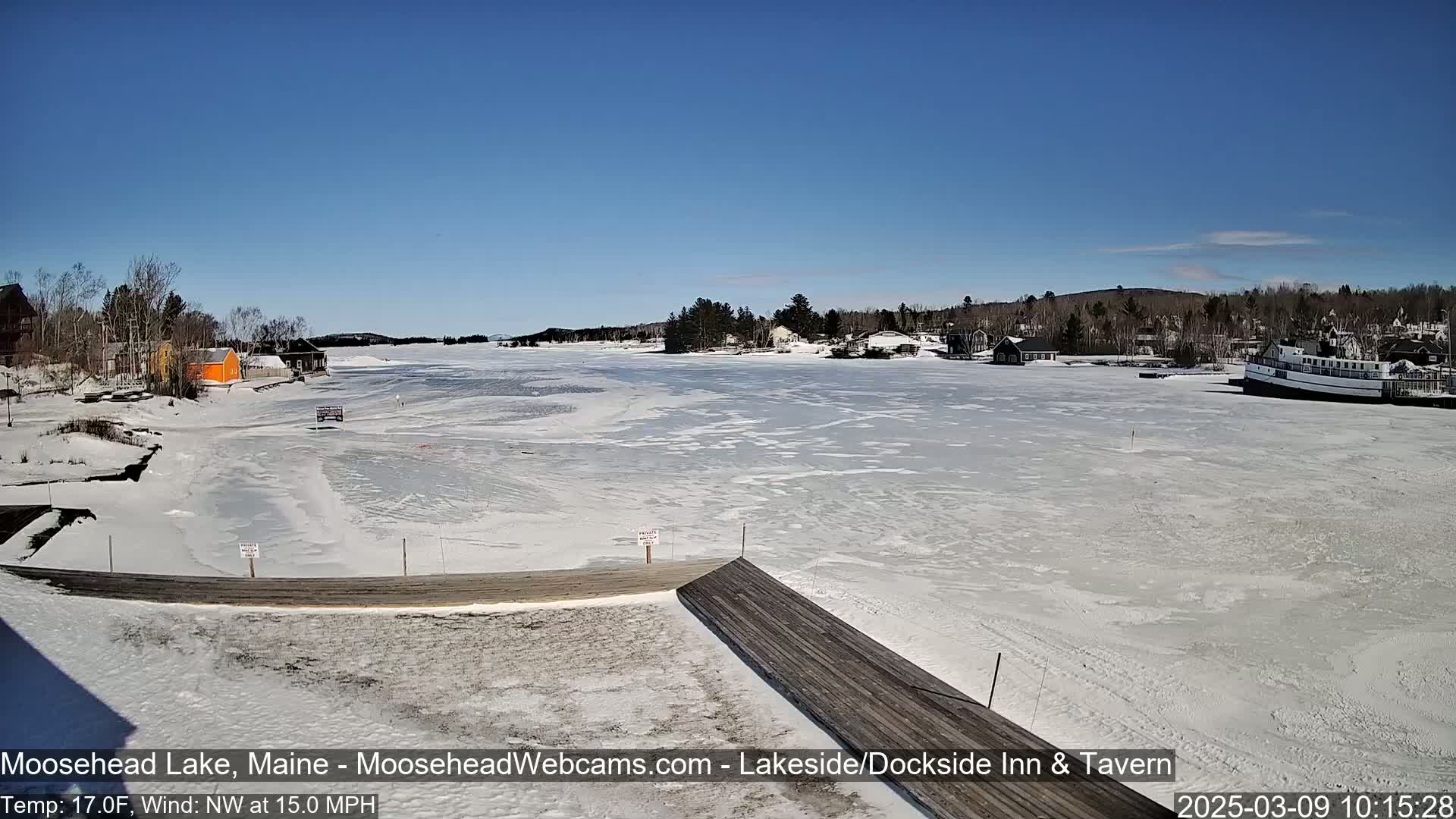 A mostly frozen lake is seen from a dock on a sunny day, with a few buildings and a boat visible in the distance.