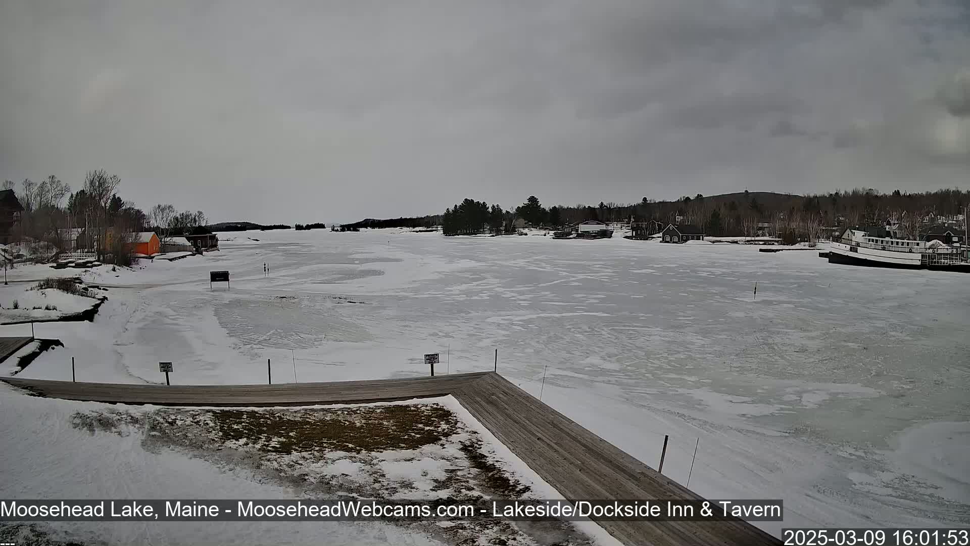 A mostly frozen lake is seen from a snowy dock, with a few buildings and trees visible along the shoreline under an overcast sky.