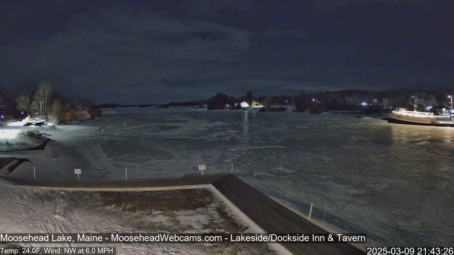 A partially frozen lake at night, viewed from a snow-covered shoreline, with a boat moored near the far shore under a dark, cloudy sky.