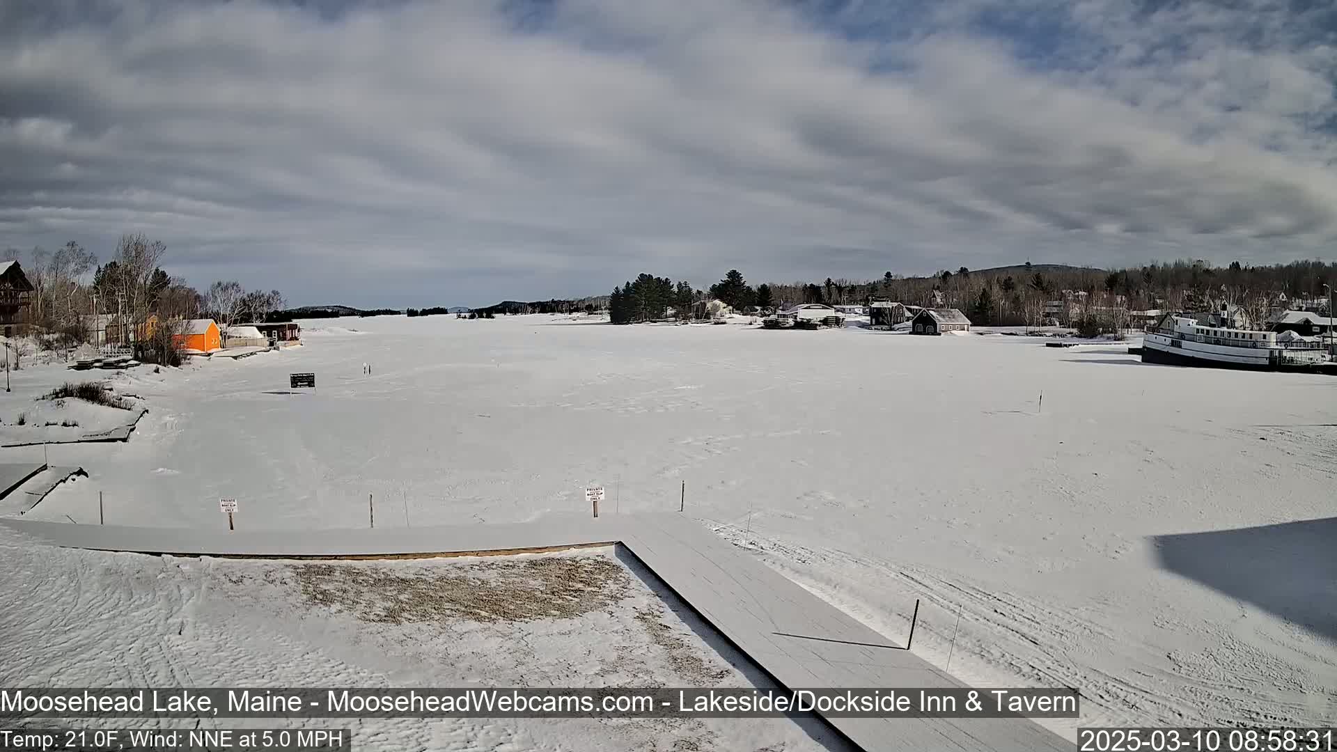 A mostly sunny day with a partly cloudy sky shows a snow-covered frozen lake with buildings and a boat in the distance.