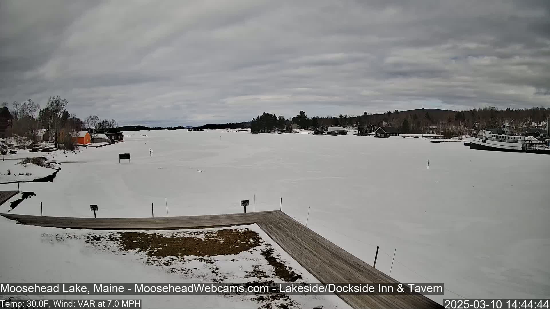 A frozen lake, under a cloudy sky, features a snow-covered shoreline with several buildings and a docked boat.
