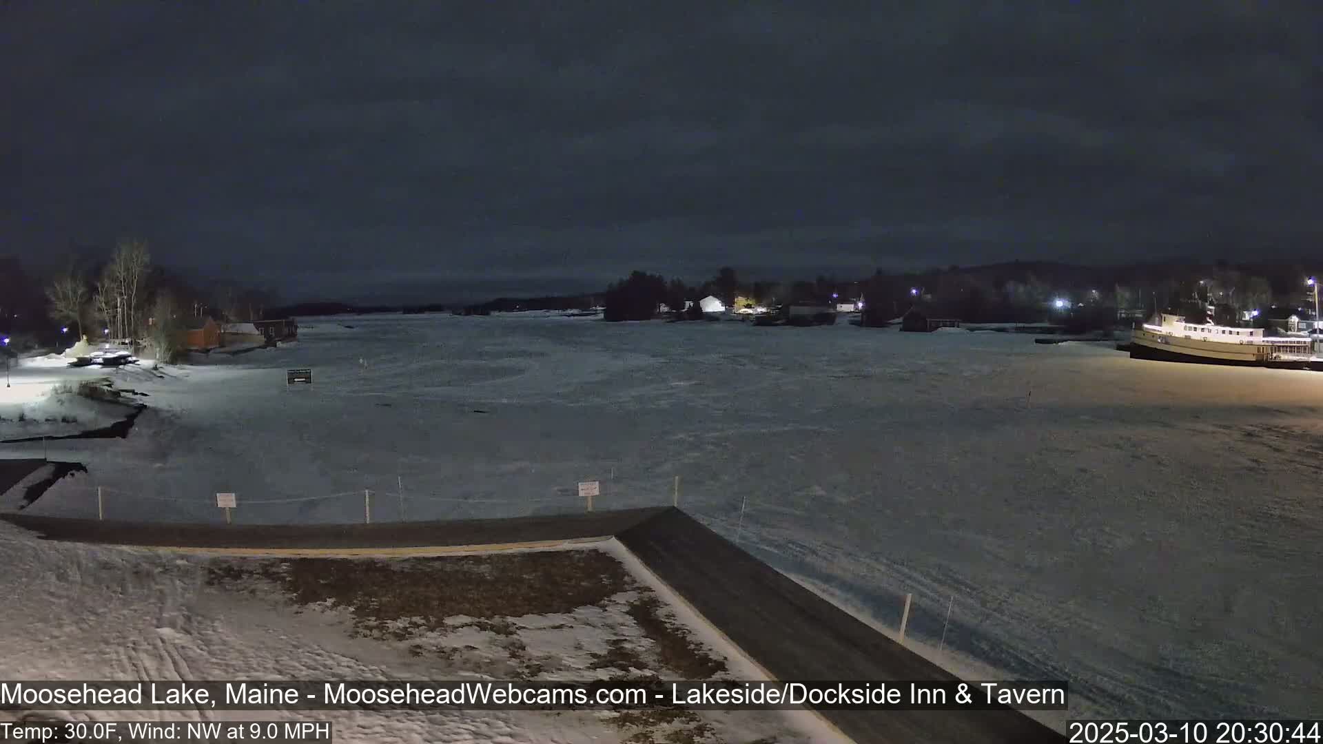 A frozen lake at night is seen from a lakeside vantage point, with a few buildings and a boat visible in the distance under a dark sky.