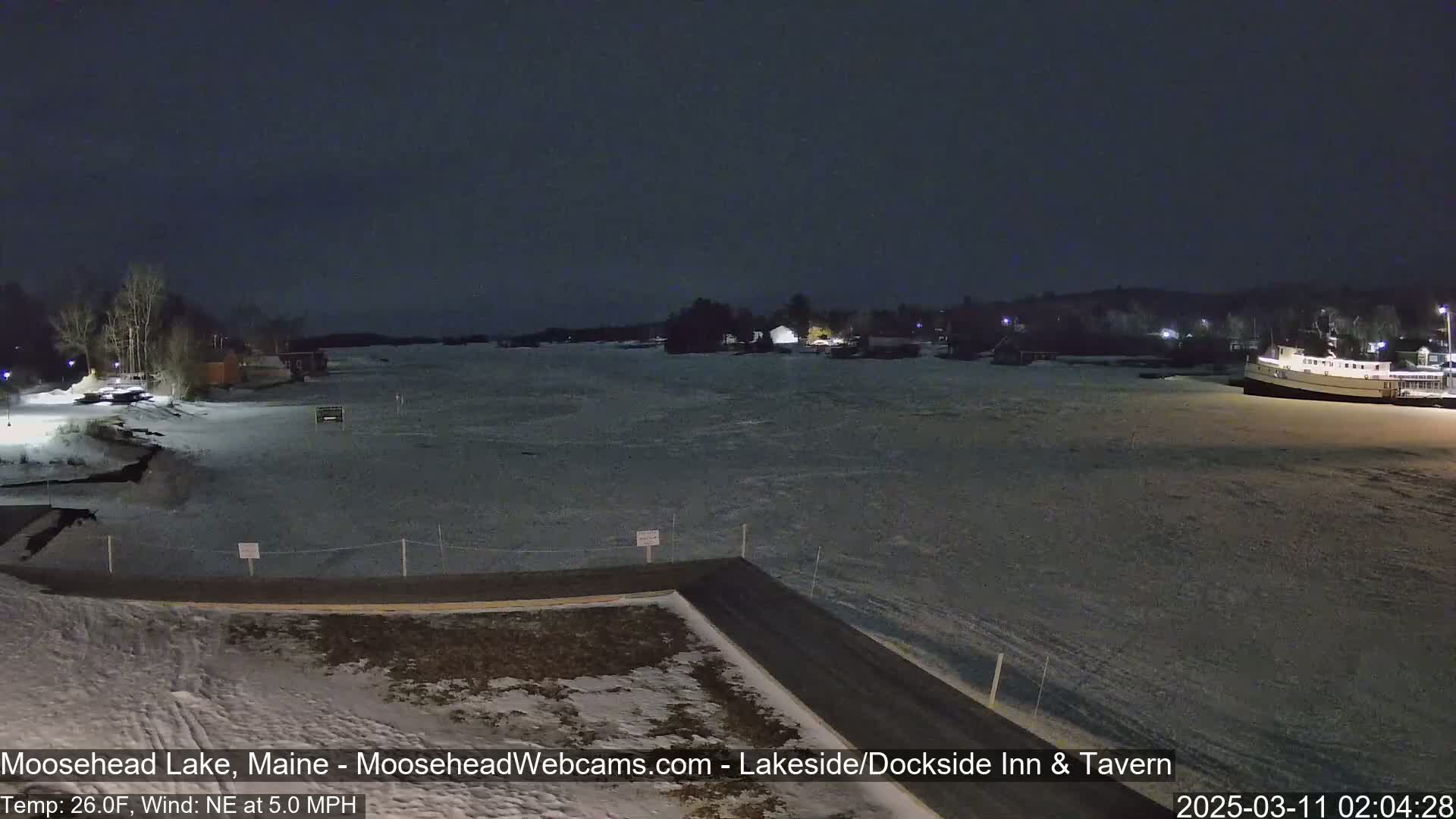 A mostly frozen lake is seen at night under a dark sky, with a few buildings and a boat visible along the shore.