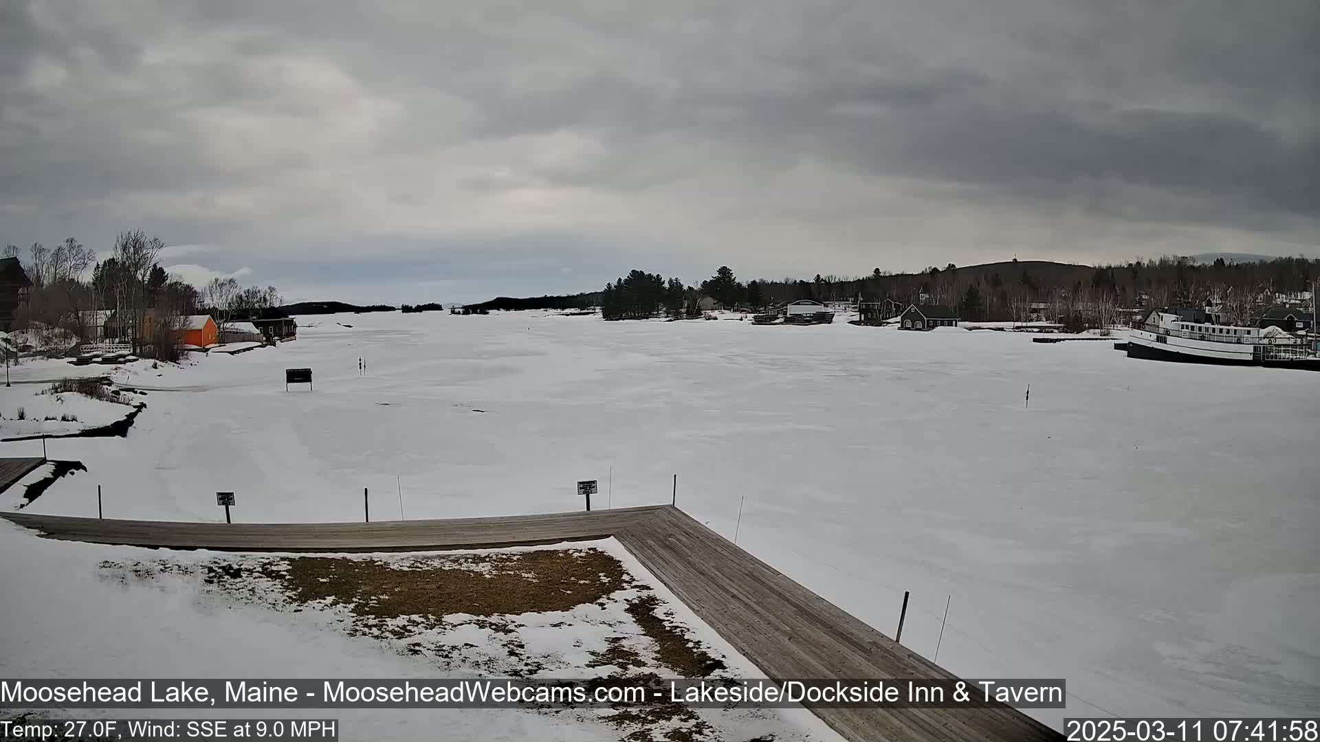 A mostly frozen lake is seen from a wooden dock, with a few buildings and boats visible along the shoreline under an overcast sky.