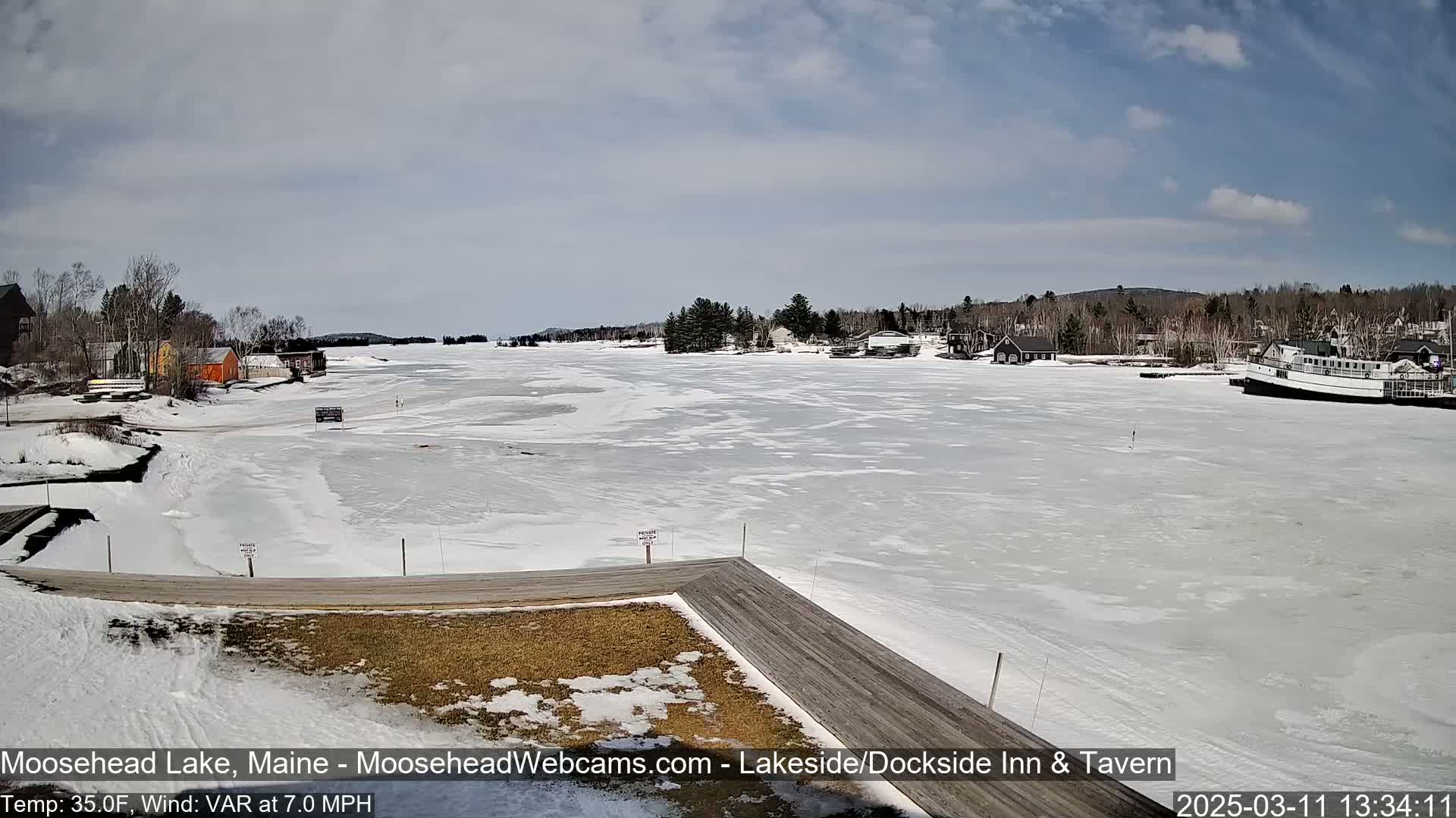 A partially frozen lake is seen from a wooden dock, with buildings and trees lining the shore under a partly cloudy sky.