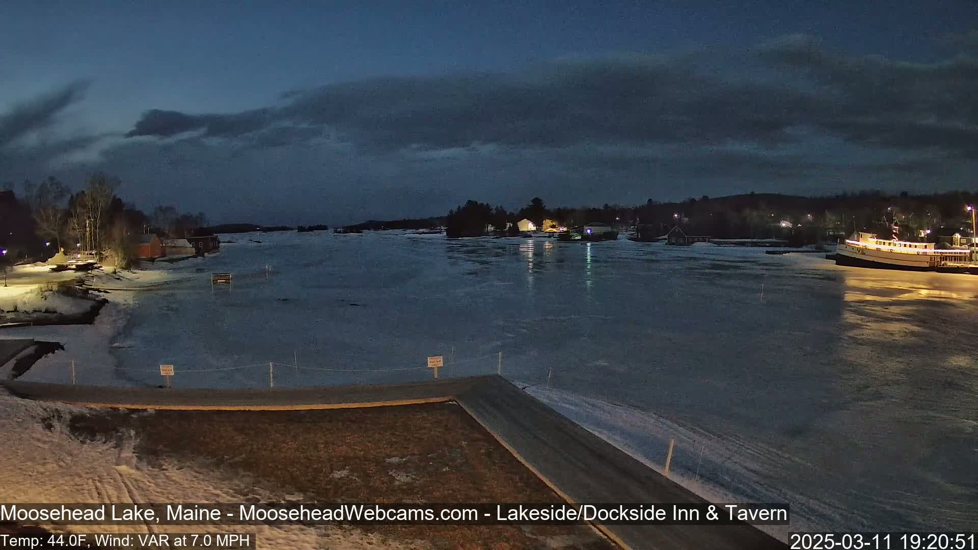 A partially frozen lake at dusk is seen from a lakeside road, with buildings and a boat visible in the distance under a cloudy sky.