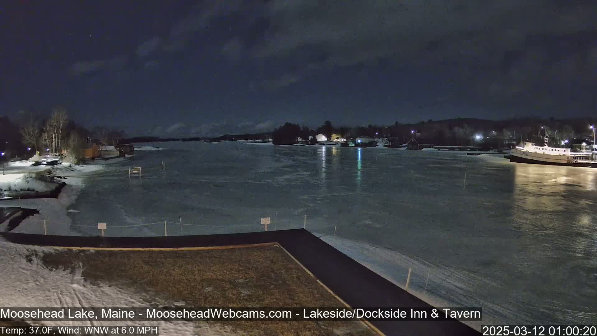 A mostly frozen lake at night, under a dark sky with a few clouds, is seen from a lakeside vantage point, showing some buildings and a boat on the far shore.