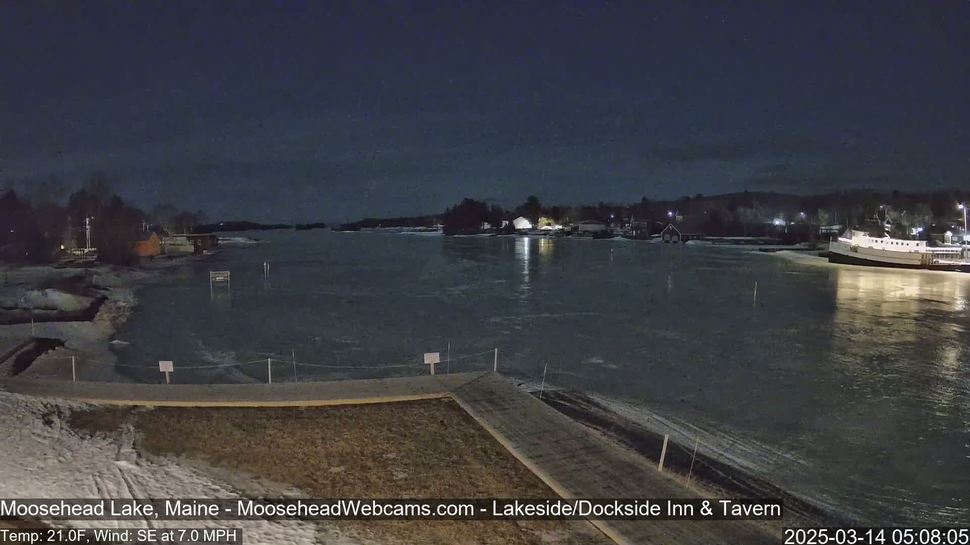 A nighttime view of a partially frozen lake shows a shoreline with buildings and a docked boat under a dark sky with a southeast wind.