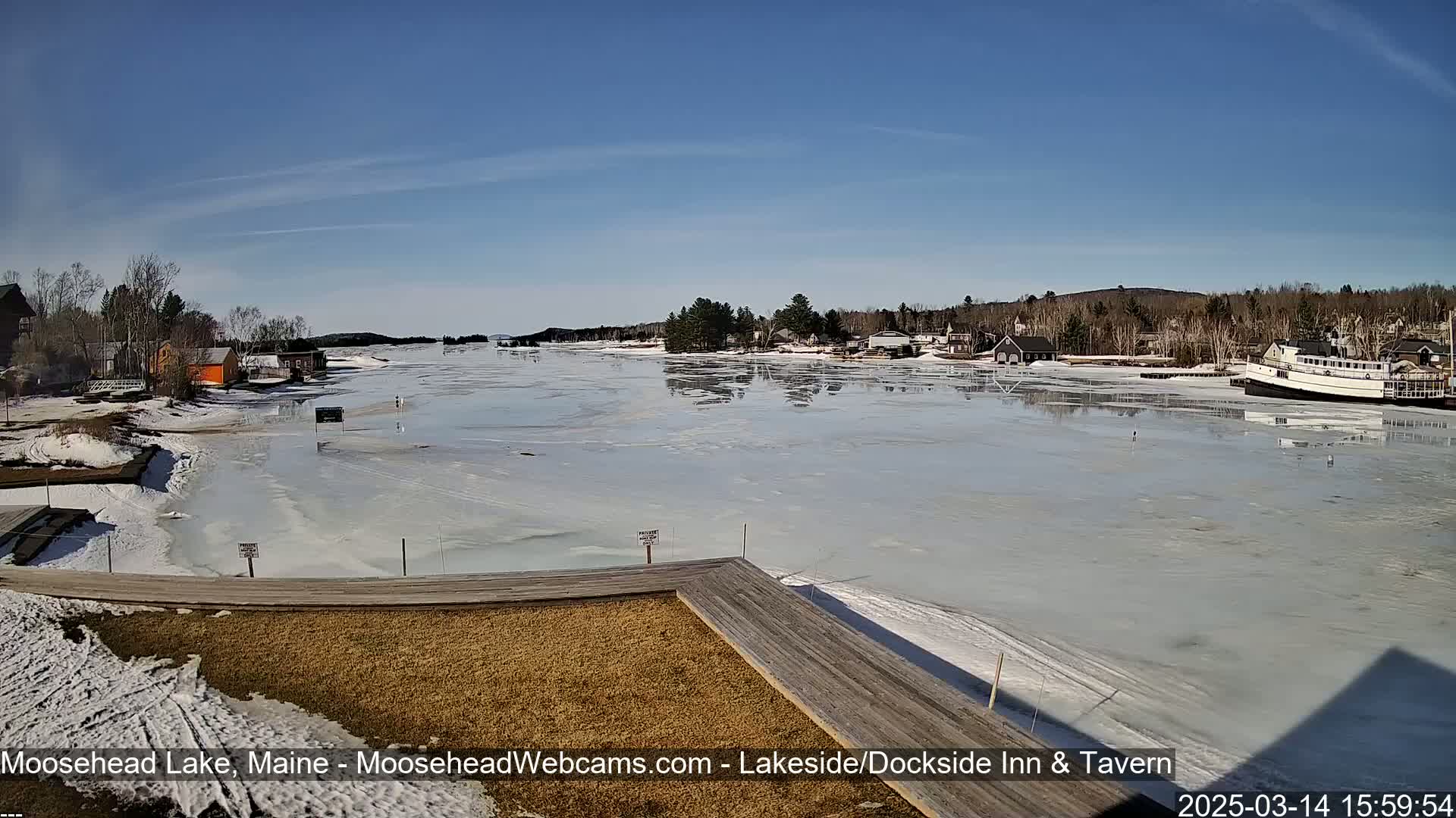 A partially frozen lake is seen from a wooden dock on a sunny day, with buildings and trees lining the shore.