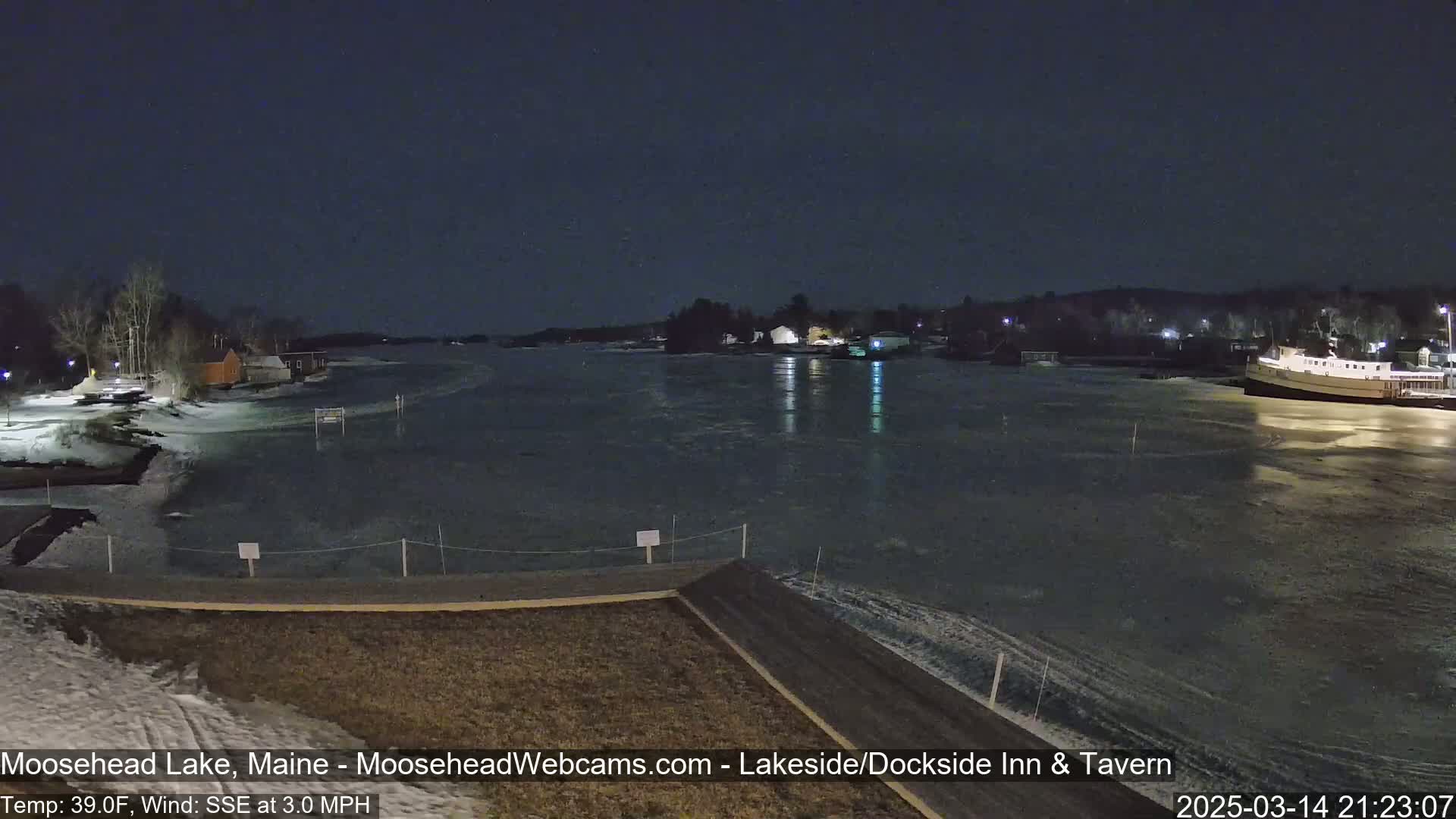 A partly ice-covered lake at night is viewed from a lakeside vantage point, with buildings and a boat visible in the distance under a dark sky.