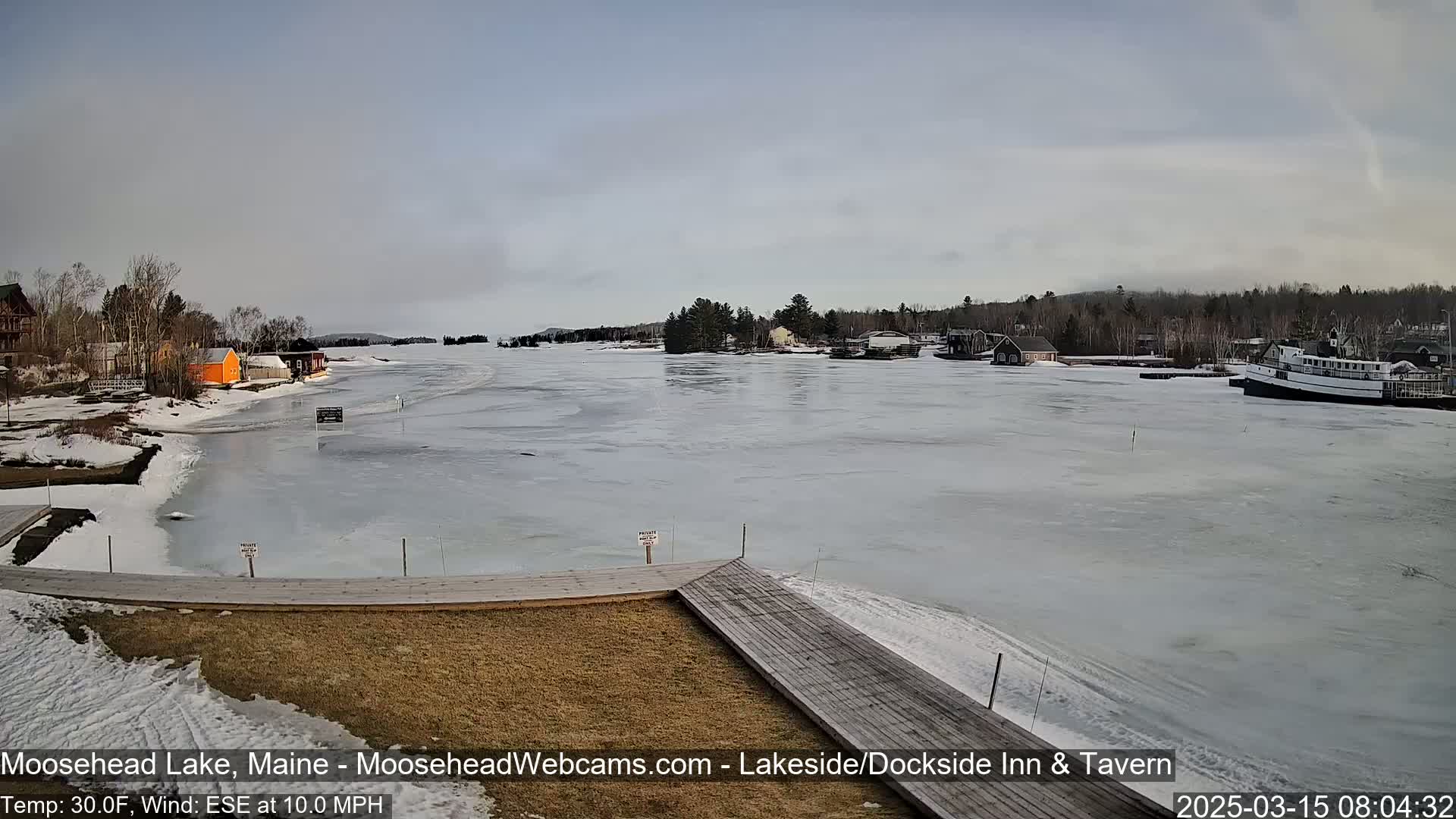 A mostly frozen lake with a few buildings and a boat visible on the shore, under an overcast sky with a 10 mph wind.