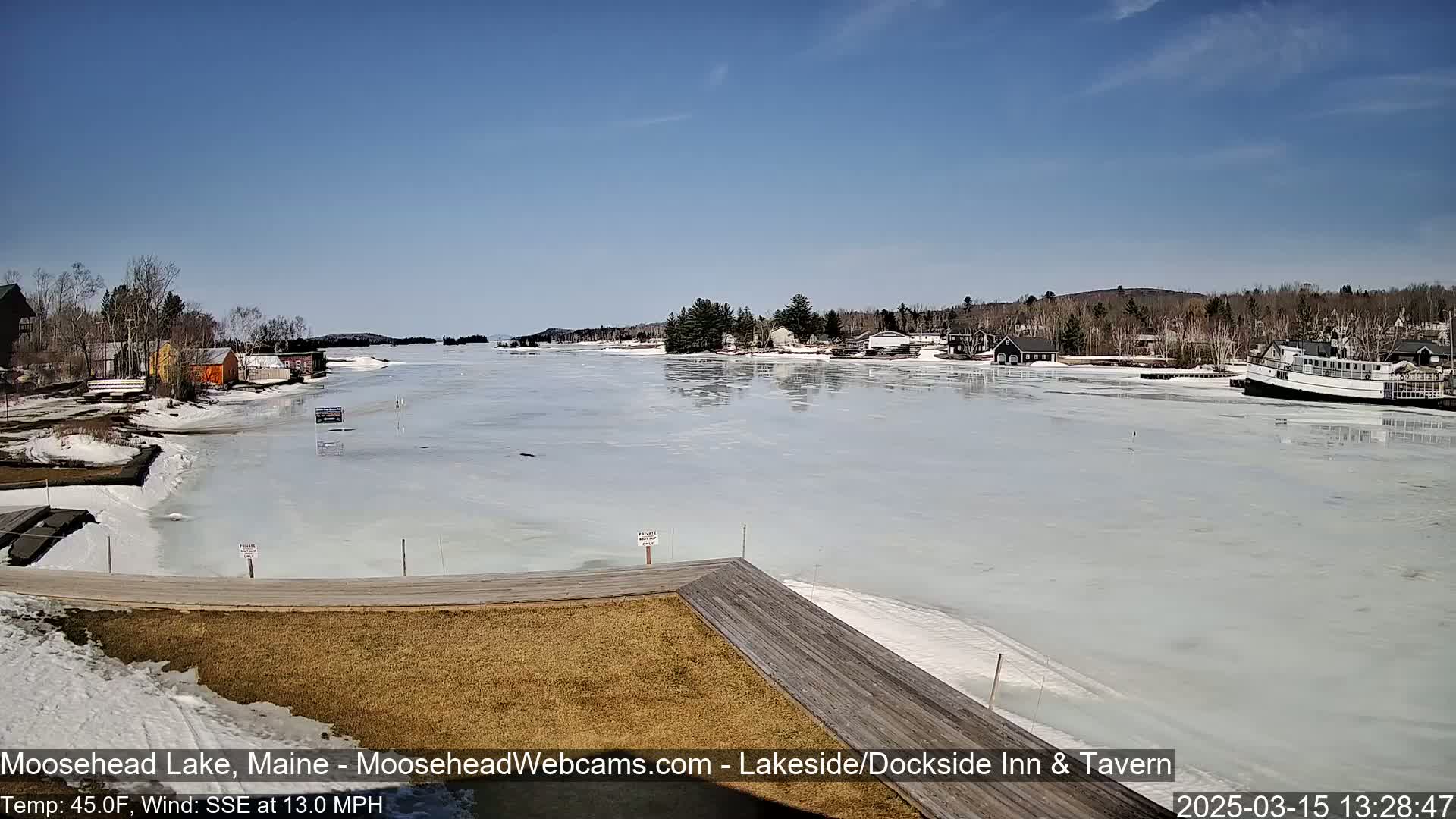 A partially frozen lake stretches out from a wooden dock, under a clear blue sky, with buildings and trees lining the shore.