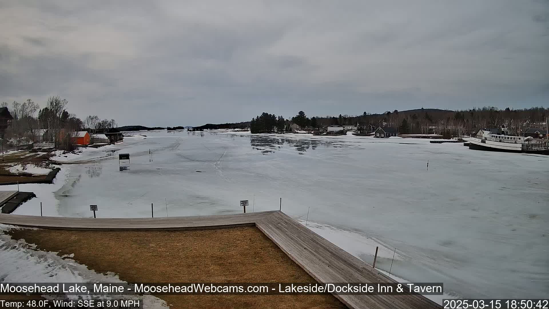 A partially frozen lake is viewed from a wooden dock, with buildings and trees lining the shore under an overcast sky.
